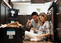 A couple is seated on the floor of a kitchen surrounded by packing boxes labeled 'Hive Boxx'. They are smiling and appear to be enjoying unpacking or eating together. The kitchen has dark wooden cabinets and a white countertop with a potted plant visible in the background.