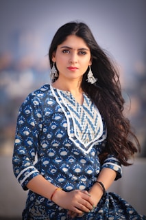 A woman with long dark hair is wearing a blue patterned traditional outfit with intricate white designs. She is accessorized with silver earrings and a small red bindi on her forehead. Her hands are clasped in front of her.
