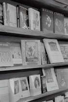 A bookshelf is filled with a variety of books, most of which have distinct covers featuring text and images. Several books are aligned upright on the top shelf, displaying a range of titles and cover designs. The books on the lower shelves are arranged in an overlapping manner, showcasing historical and cultural themes.