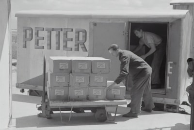 Two individuals are unloading or loading a stack of boxes from the back of a trailer or truck. The trailer has the word 'PETTER' written on its side. One person stands inside the trailer while the other is outside arranging the boxes on a cart.