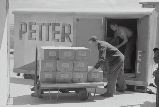 Two individuals are unloading or loading a stack of boxes from the back of a trailer or truck. The trailer has the word 'PETTER' written on its side. One person stands inside the trailer while the other is outside arranging the boxes on a cart.