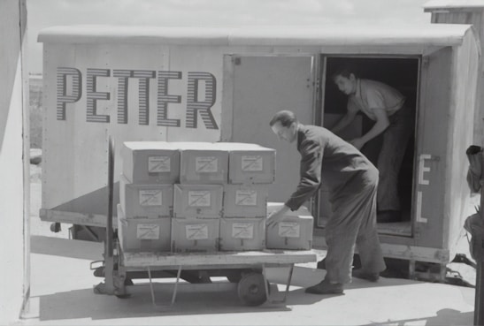 Two individuals are unloading or loading a stack of boxes from the back of a trailer or truck. The trailer has the word 'PETTER' written on its side. One person stands inside the trailer while the other is outside arranging the boxes on a cart.
