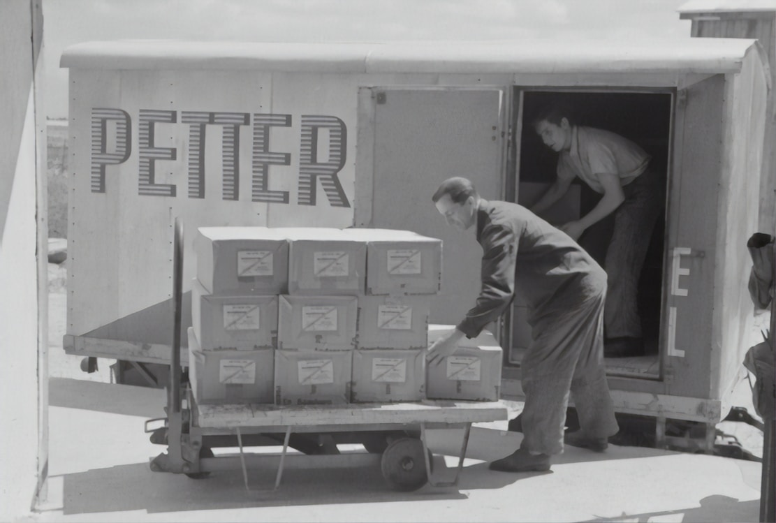 Two individuals are unloading or loading a stack of boxes from the back of a trailer or truck. The trailer has the word 'PETTER' written on its side. One person stands inside the trailer while the other is outside arranging the boxes on a cart.