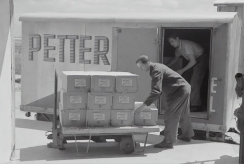 Two individuals are unloading or loading a stack of boxes from the back of a trailer or truck. The trailer has the word 'PETTER' written on its side. One person stands inside the trailer while the other is outside arranging the boxes on a cart.