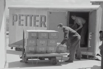 Two individuals are unloading or loading a stack of boxes from the back of a trailer or truck. The trailer has the word 'PETTER' written on its side. One person stands inside the trailer while the other is outside arranging the boxes on a cart.