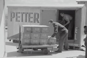 Two individuals are unloading or loading a stack of boxes from the back of a trailer or truck. The trailer has the word 'PETTER' written on its side. One person stands inside the trailer while the other is outside arranging the boxes on a cart.