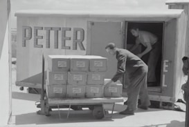 Two individuals are unloading or loading a stack of boxes from the back of a trailer or truck. The trailer has the word 'PETTER' written on its side. One person stands inside the trailer while the other is outside arranging the boxes on a cart.