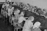 A group of women working together at a long table inside an industrial setting. They are wearing work attire, including headscarves and aprons, and appear to be sorting or processing a material laid out on the table. The room has tiled floors and walls, suggesting a factory environment.