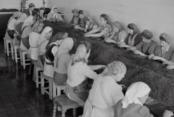 A group of women working together at a long table inside an industrial setting. They are wearing work attire, including headscarves and aprons, and appear to be sorting or processing a material laid out on the table. The room has tiled floors and walls, suggesting a factory environment.