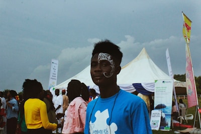 A person with face paint stands in the foreground wearing a blue shirt with a white puzzle piece design. In the background, several people are gathered around a large white tent, with various banners and promotional signs visible.
