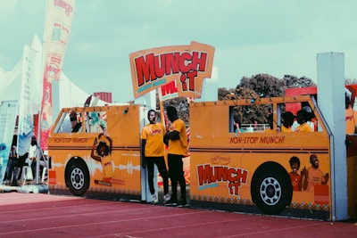 Several people are standing behind a large, bright yellow cardboard cutout of a bus that promotes 'Munch It' crunchy snacks. The cutout features vibrant graphics and slogans, with a prominent display of the snack's branding. The scene is set outdoors, possibly at an event or fairground, with flags and tents visible in the background.