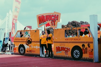 Several people are standing behind a large, bright yellow cardboard cutout of a bus that promotes 'Munch It' crunchy snacks. The cutout features vibrant graphics and slogans, with a prominent display of the snack's branding. The scene is set outdoors, possibly at an event or fairground, with flags and tents visible in the background.