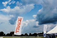 A large outdoor space displays a tall white flag with 'TRACE' written in bold red letters. In the background, a dramatic sky filled with thick, dark clouds looms over a wide expanse of green land bordered by trees. A tent with a promotional banner that reads 'It Pays To Save With Fidelity' is visible, suggesting a sponsored event or gathering. The overall scene is open and expansive, indicative of a field or park setting.