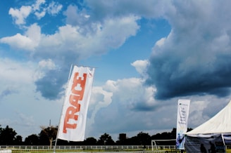 A large outdoor space displays a tall white flag with 'TRACE' written in bold red letters. In the background, a dramatic sky filled with thick, dark clouds looms over a wide expanse of green land bordered by trees. A tent with a promotional banner that reads 'It Pays To Save With Fidelity' is visible, suggesting a sponsored event or gathering. The overall scene is open and expansive, indicative of a field or park setting.
