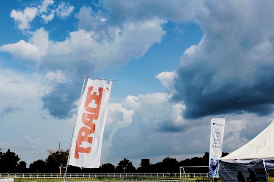 A large outdoor space displays a tall white flag with 'TRACE' written in bold red letters. In the background, a dramatic sky filled with thick, dark clouds looms over a wide expanse of green land bordered by trees. A tent with a promotional banner that reads 'It Pays To Save With Fidelity' is visible, suggesting a sponsored event or gathering. The overall scene is open and expansive, indicative of a field or park setting.
