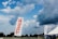 A large outdoor space displays a tall white flag with 'TRACE' written in bold red letters. In the background, a dramatic sky filled with thick, dark clouds looms over a wide expanse of green land bordered by trees. A tent with a promotional banner that reads 'It Pays To Save With Fidelity' is visible, suggesting a sponsored event or gathering. The overall scene is open and expansive, indicative of a field or park setting.