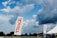 A large outdoor space displays a tall white flag with 'TRACE' written in bold red letters. In the background, a dramatic sky filled with thick, dark clouds looms over a wide expanse of green land bordered by trees. A tent with a promotional banner that reads 'It Pays To Save With Fidelity' is visible, suggesting a sponsored event or gathering. The overall scene is open and expansive, indicative of a field or park setting.