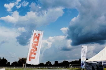 A large outdoor space displays a tall white flag with 'TRACE' written in bold red letters. In the background, a dramatic sky filled with thick, dark clouds looms over a wide expanse of green land bordered by trees. A tent with a promotional banner that reads 'It Pays To Save With Fidelity' is visible, suggesting a sponsored event or gathering. The overall scene is open and expansive, indicative of a field or park setting.