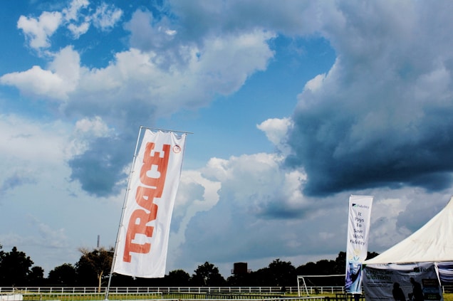 A large outdoor space displays a tall white flag with 'TRACE' written in bold red letters. In the background, a dramatic sky filled with thick, dark clouds looms over a wide expanse of green land bordered by trees. A tent with a promotional banner that reads 'It Pays To Save With Fidelity' is visible, suggesting a sponsored event or gathering. The overall scene is open and expansive, indicative of a field or park setting.