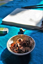 A ceramic bowl containing pencil shavings sits on a vibrant blue fabric surface. Nearby, there is an eraser and a set of pencils, with a sketch pad partially visible, suggesting a workspace for drawing or sketching.