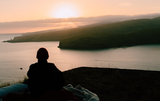 man in black jacket sitting on green grass near body of water during daytime in Godley Head Road New Zealand