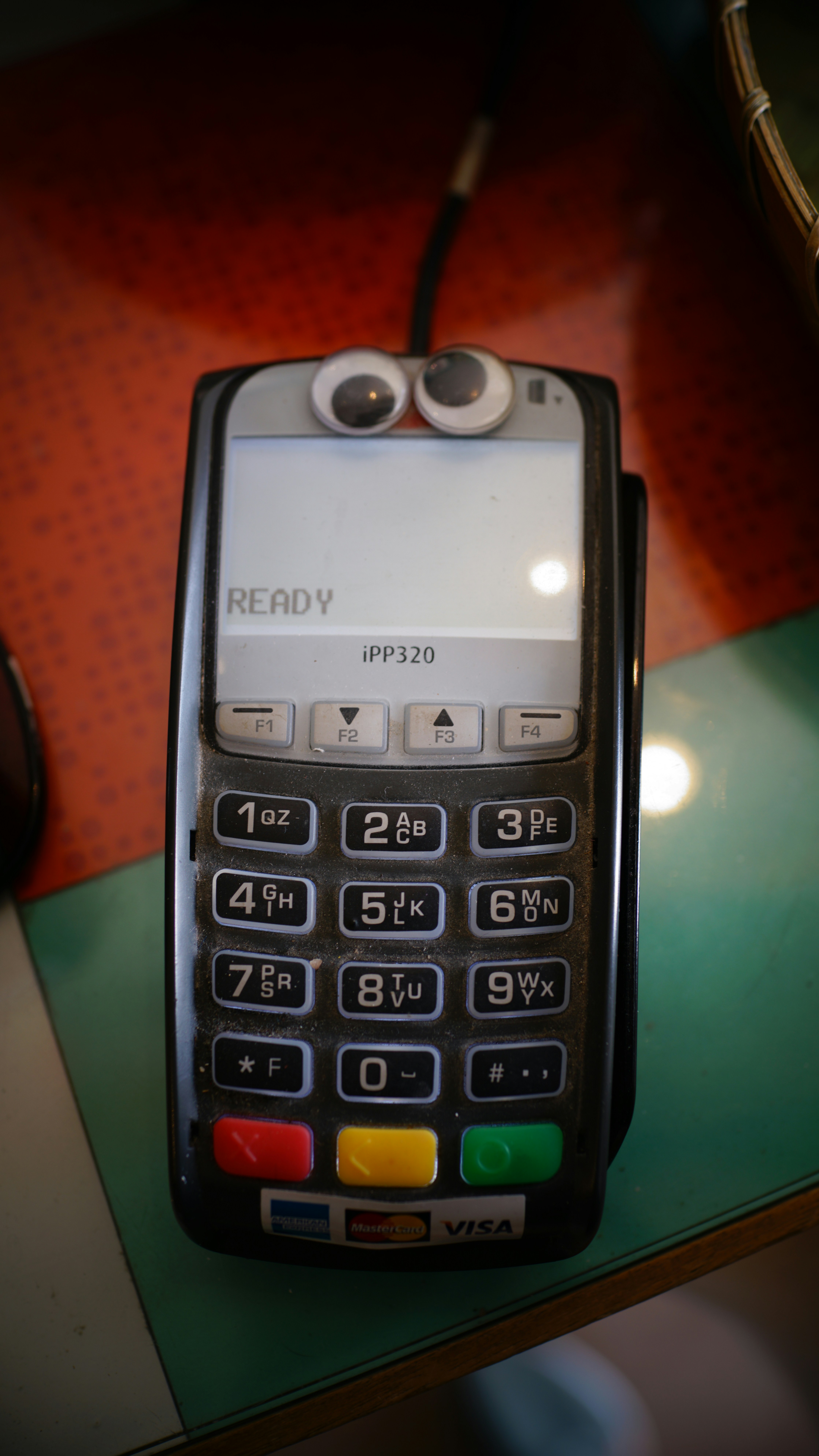 A payment card reader with a keypad and a display screen on top. Googly eyes are attached above the screen, giving it a playful appearance. The device has several buttons numbered from 0 to 9, along with function keys and color-coded buttons at the bottom. It displays the word 'READY' on the screen.