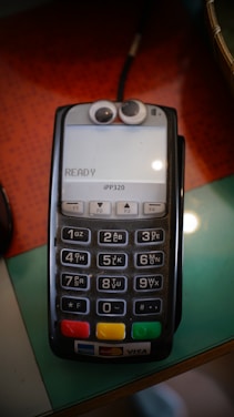 A payment card reader with a keypad and a display screen on top. Googly eyes are attached above the screen, giving it a playful appearance. The device has several buttons numbered from 0 to 9, along with function keys and color-coded buttons at the bottom. It displays the word 'READY' on the screen.