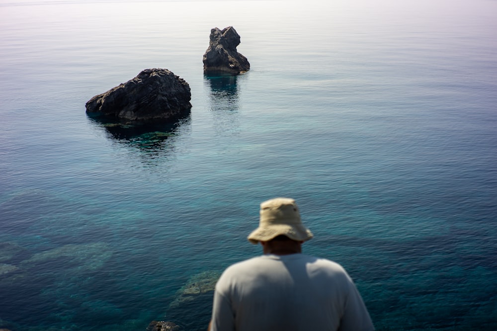 man in white shirt wearing brown hat sitting on rock in front of body of water