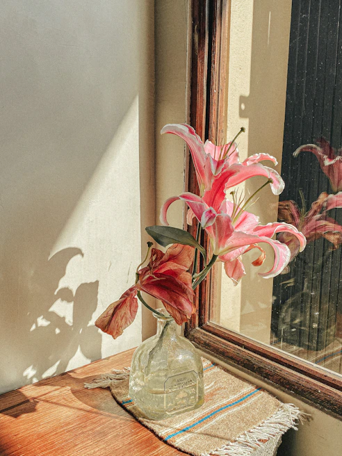 A tall glass vase filled with fresh white lilies, placed on a sunlit windowsill