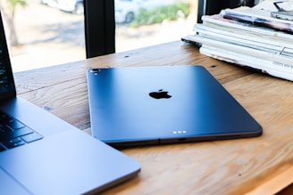 A sleek Apple iPad lies on a wooden table next to a partially visible MacBook. Nearby, there is a stack of magazines or newspapers on the right side. Sunlight filters in through windows, casting soft shadows.