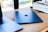 A sleek Apple iPad lies on a wooden table next to a partially visible MacBook. Nearby, there is a stack of magazines or newspapers on the right side. Sunlight filters in through windows, casting soft shadows.