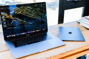 A laptop with an open screen displaying a nature-themed wallpaper is placed on a wooden desk next to a closed tablet. There are stacks of magazines or papers on the right side and the setting appears to be near a window with natural light coming in.