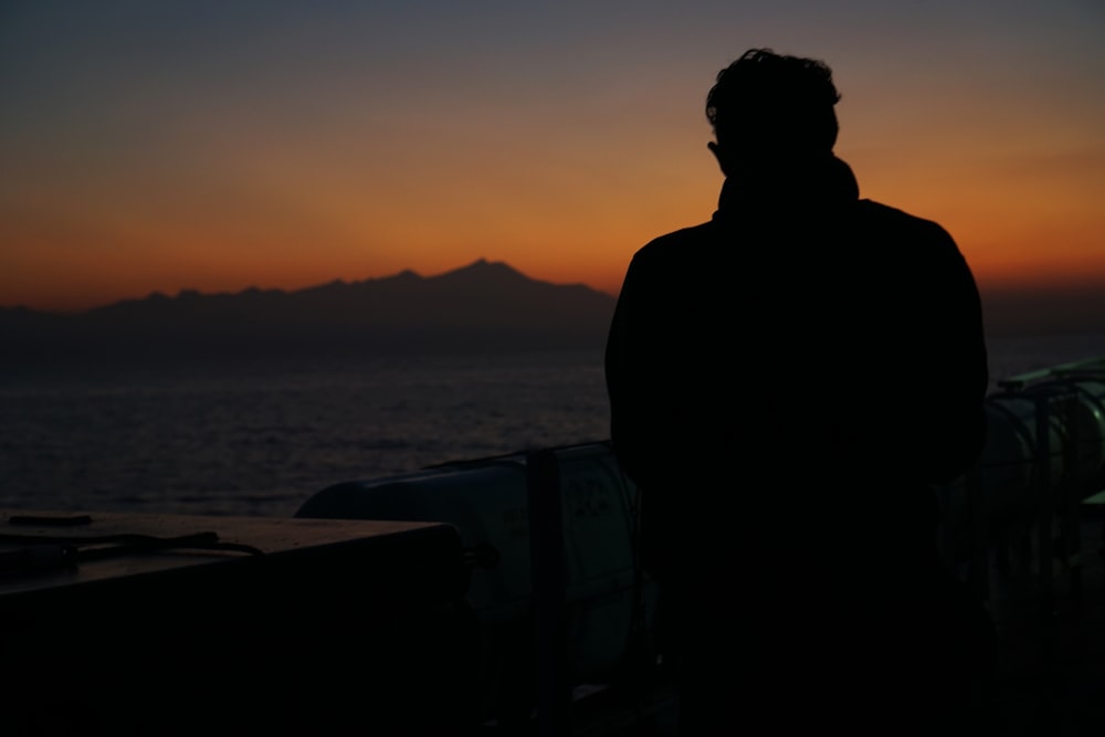 silhouette of man standing near body of water during sunset