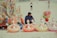 A person sits on a sandy beach surrounded by inflatable duck rings and assorted beach toys on display. In the background, there are two people walking along the beach. Colorful items hang from a display stand, and a cluster of vibrant balloons is attached to it.