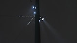 A dark night sky with a tall street light pole illuminated by multiple bright spotlights. The lights create a misty effect as they shine through the rain or fog, highlighting raindrops or particles in the air. The pole appears robust, with multiple clamps and wires extending horizontally.