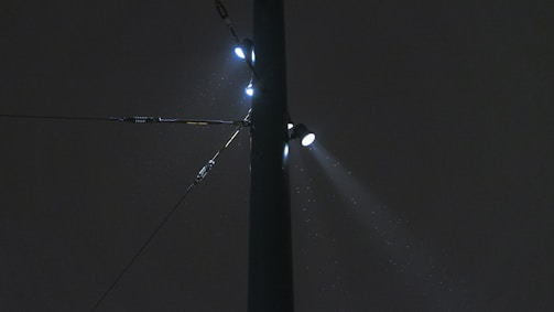 A dark night sky with a tall street light pole illuminated by multiple bright spotlights. The lights create a misty effect as they shine through the rain or fog, highlighting raindrops or particles in the air. The pole appears robust, with multiple clamps and wires extending horizontally.