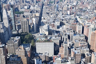 A vast urban landscape featuring numerous densely packed skyscrapers and buildings. The image is taken from an elevated perspective, showcasing the varied architecture of the city. Green patches of trees and parks are visible amidst the concrete structures, adding contrast to the scene.