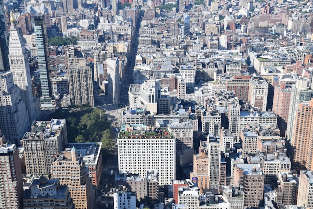 A vast urban landscape featuring numerous densely packed skyscrapers and buildings. The image is taken from an elevated perspective, showcasing the varied architecture of the city. Green patches of trees and parks are visible amidst the concrete structures, adding contrast to the scene.