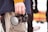 A person holding a Panasonic Lumix camera in their right hand. The individual is wearing a blue jacket and beige pants. The background is slightly blurred, with indistinct figures and a colorful object that resembles a patterned blanket.
