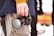 A person holding a Panasonic Lumix camera in their right hand. The individual is wearing a blue jacket and beige pants. The background is slightly blurred, with indistinct figures and a colorful object that resembles a patterned blanket.