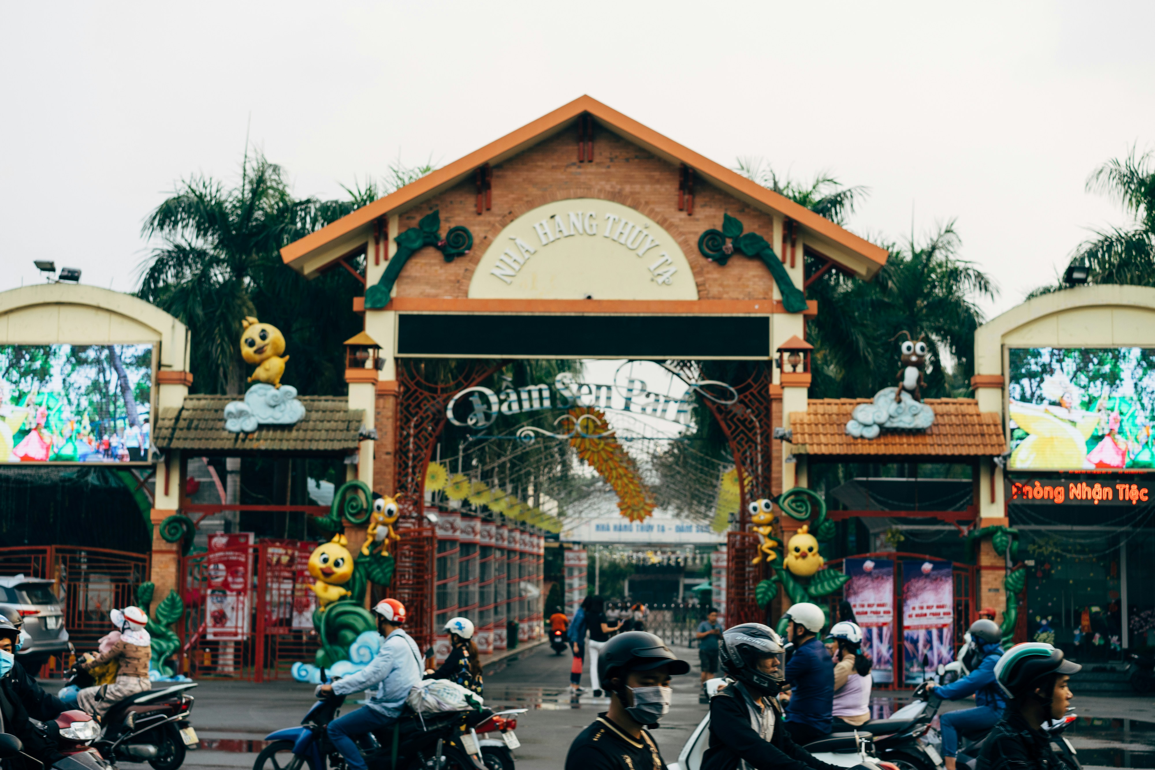 people riding motorcycle on road near brown wooden building during daytime, Entrance to the Dam Den Park in Ho Chi Minh CIty