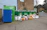 A collection of large green recycling bins are filled with overflowing bags and items, surrounded by numerous plastic bags full of recyclables on the ground. A blue donation bin labeled for clothes and shoes is nearby, situated against a brick building near a street.