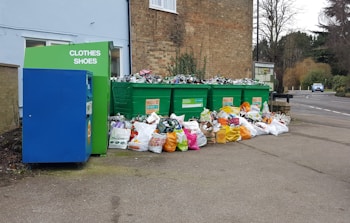 A collection of large green recycling bins are filled with overflowing bags and items, surrounded by numerous plastic bags full of recyclables on the ground. A blue donation bin labeled for clothes and shoes is nearby, situated against a brick building near a street.