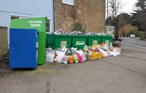 A collection of large green recycling bins are filled with overflowing bags and items, surrounded by numerous plastic bags full of recyclables on the ground. A blue donation bin labeled for clothes and shoes is nearby, situated against a brick building near a street.