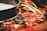 Close-up view of a hot pot containing red chili peppers and various spices in a dark, oily broth. A portion of the pot rim is visible, indicating it's a shared dish. The image captures the glossy texture and vibrant colors of the ingredients.