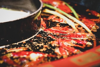 Close-up view of a hot pot containing red chili peppers and various spices in a dark, oily broth. A portion of the pot rim is visible, indicating it's a shared dish. The image captures the glossy texture and vibrant colors of the ingredients.