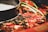 Close-up view of a hot pot containing red chili peppers and various spices in a dark, oily broth. A portion of the pot rim is visible, indicating it's a shared dish. The image captures the glossy texture and vibrant colors of the ingredients.
