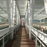 A modern pedestrian bridge with white metal beams and wooden flooring. The bridge is enclosed with glass panels, creating a tunnel-like effect. Sunlight casts geometric shadows on the floor, leading to an open area in the distance where two people are visible. A historic building with red brick and decorative elements is partially visible in the background.