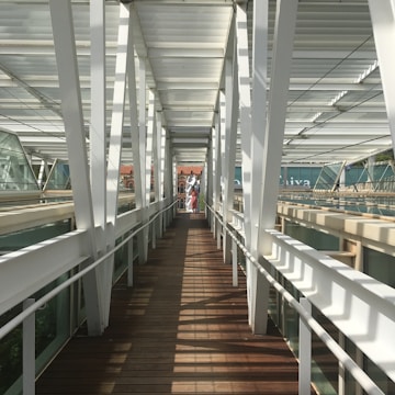 A modern pedestrian bridge with white metal beams and wooden flooring. The bridge is enclosed with glass panels, creating a tunnel-like effect. Sunlight casts geometric shadows on the floor, leading to an open area in the distance where two people are visible. A historic building with red brick and decorative elements is partially visible in the background.