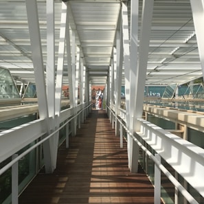 A modern pedestrian bridge with white metal beams and wooden flooring. The bridge is enclosed with glass panels, creating a tunnel-like effect. Sunlight casts geometric shadows on the floor, leading to an open area in the distance where two people are visible. A historic building with red brick and decorative elements is partially visible in the background.